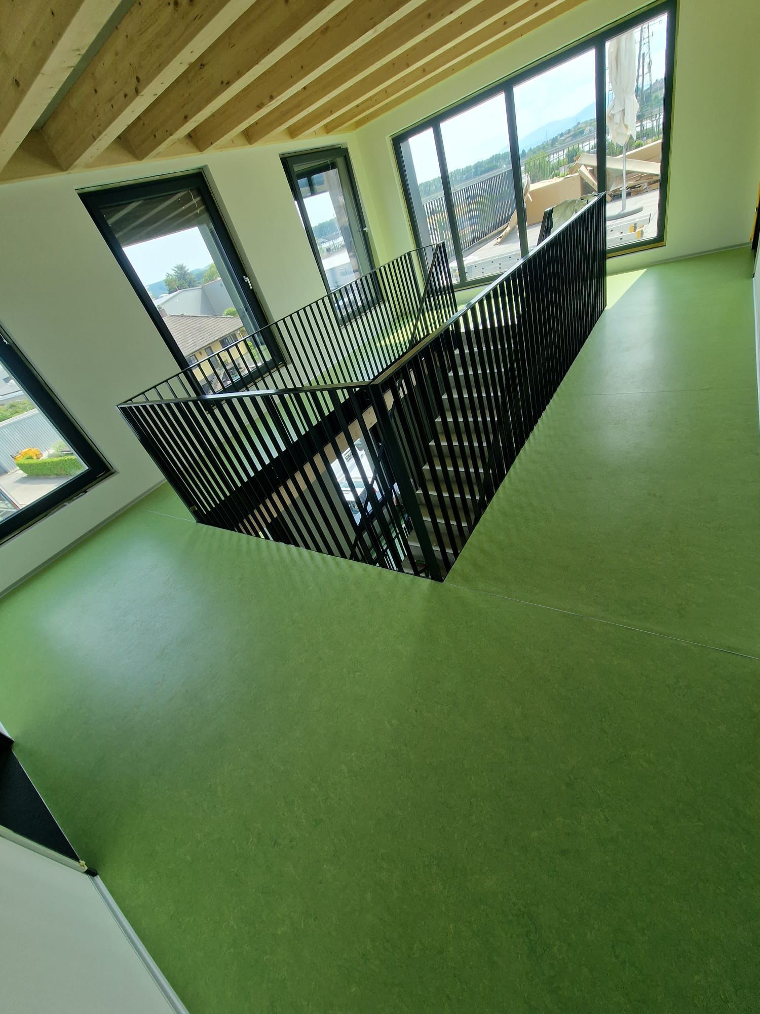 Vue intérieure d'une pièce avec sol vert, poutres apparentes au plafond, grandes fenêtres et rampe d'escalier en métal noir.