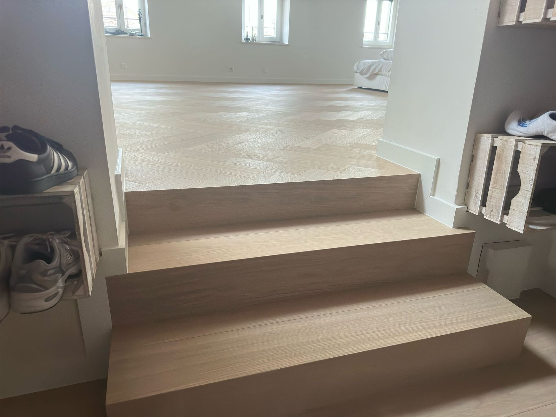 Deux marches en bois clair mènent à une pièce lumineuse et ouverte, aux murs blancs et percée de trois fenêtres. Des caisses en bois font office d'étagères.