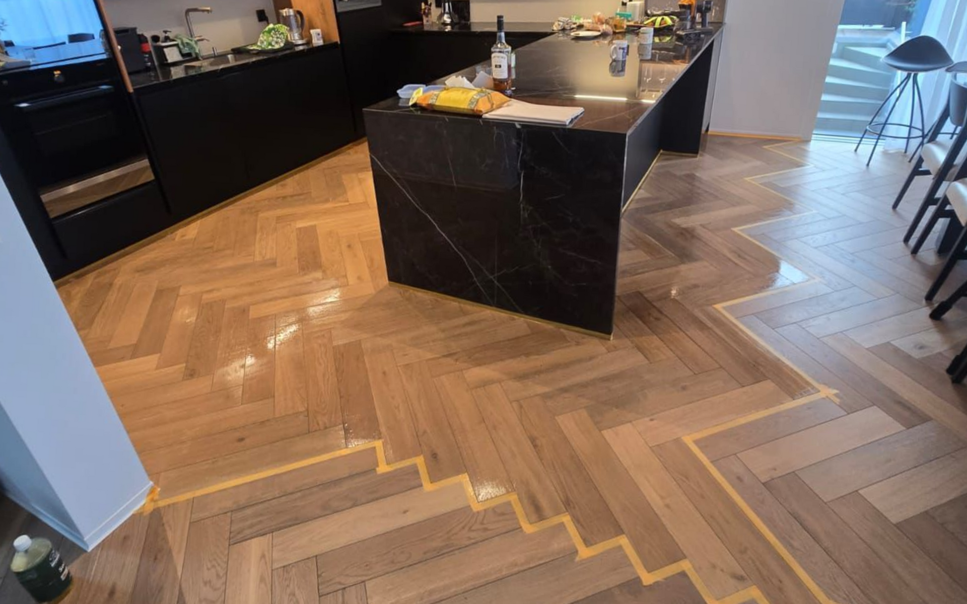 Une cuisine moderne présente un parquet à chevrons avec une ligne de délimitation distincte marquée par du ruban adhésif de peintre jaune.