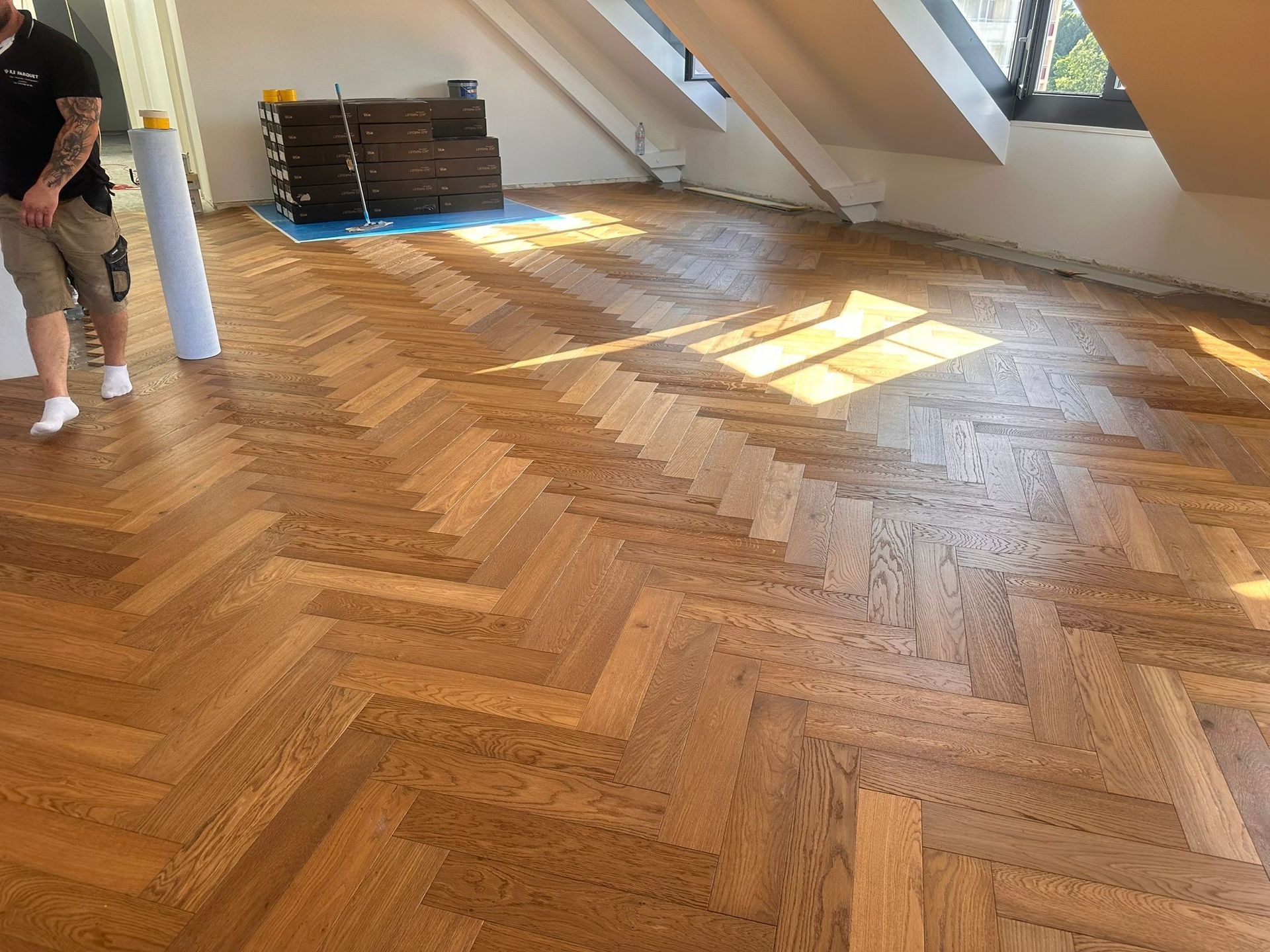Une personne se tient debout dans une pièce baignée de soleil, en train d'installer un parquet en chêne à chevrons.