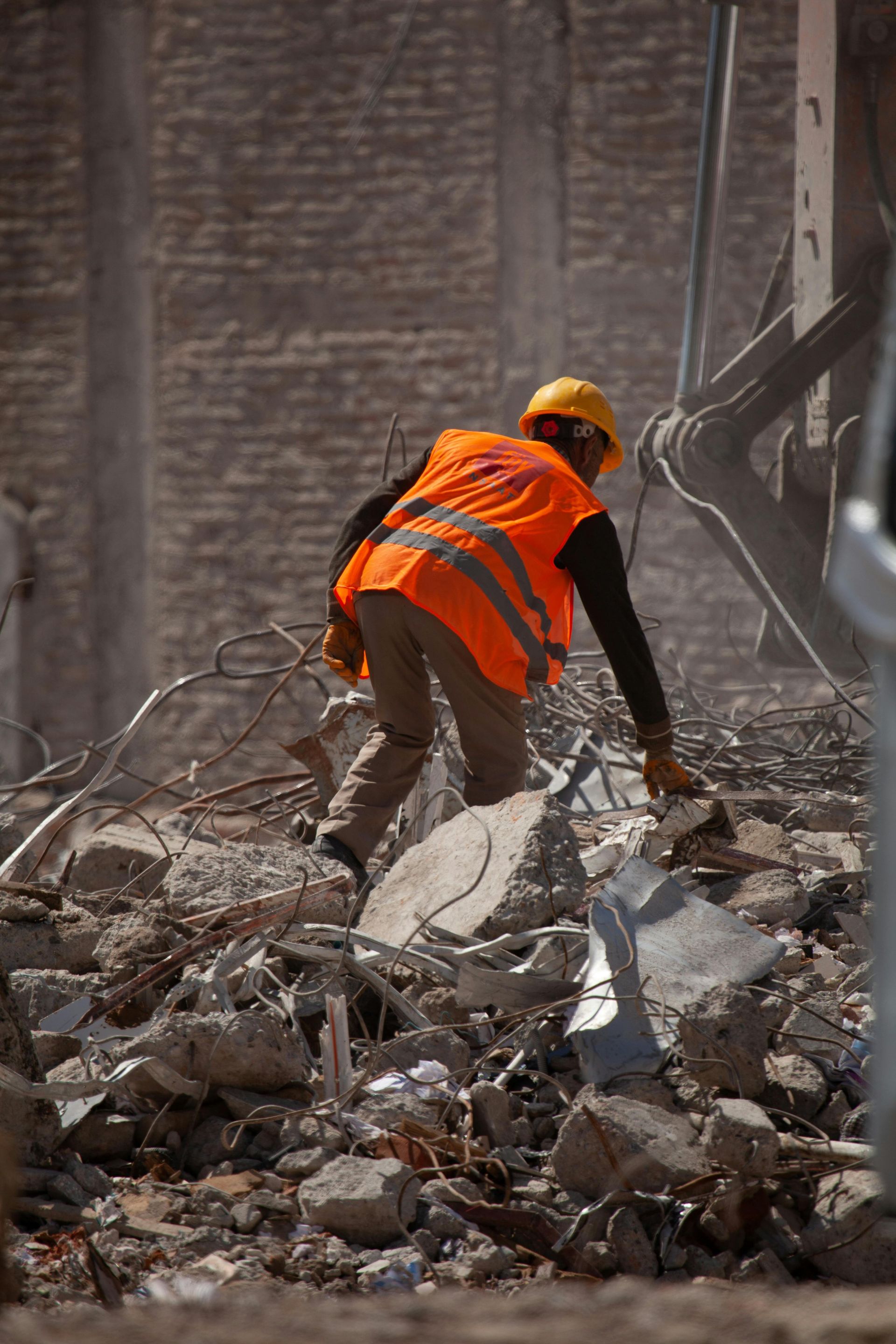 Construction worker in orange vest and hard hat searches debris at a demolition site.