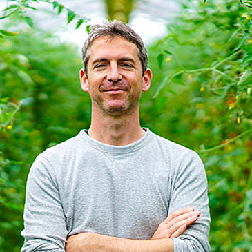 A man in a grey sweater is standing with his arms crossed in front of a bush.