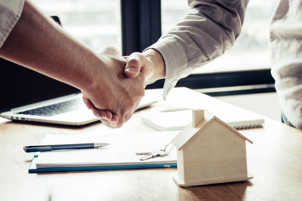 Two People Are Shaking Hands Over A Table With A Model House In The Background — Spencer Lawyers In Gosford, NSW