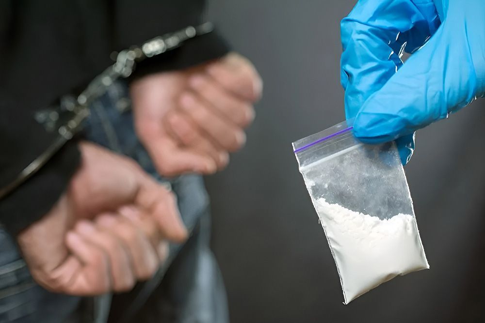 Handcuffed Person, Hand in Gloves Holding a Bag of White Powder, Likely Drugs