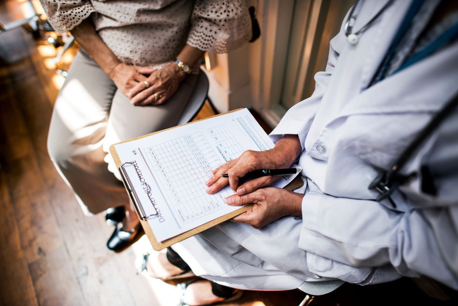 Doctor in lab coat holds clipboard, consulting with seated patient in beige attire.