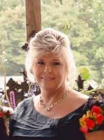 Woman with blonde hair wearing a necklace, smiling outdoors, flowers and greenery in the background.