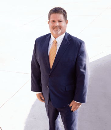 Man in navy suit and gold tie stands outdoors, smiling slightly. Bright sunlight.