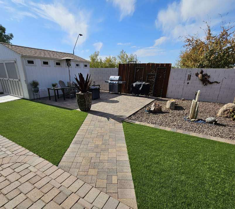 A backyard with a brick walkway leading to a patio with a grill.