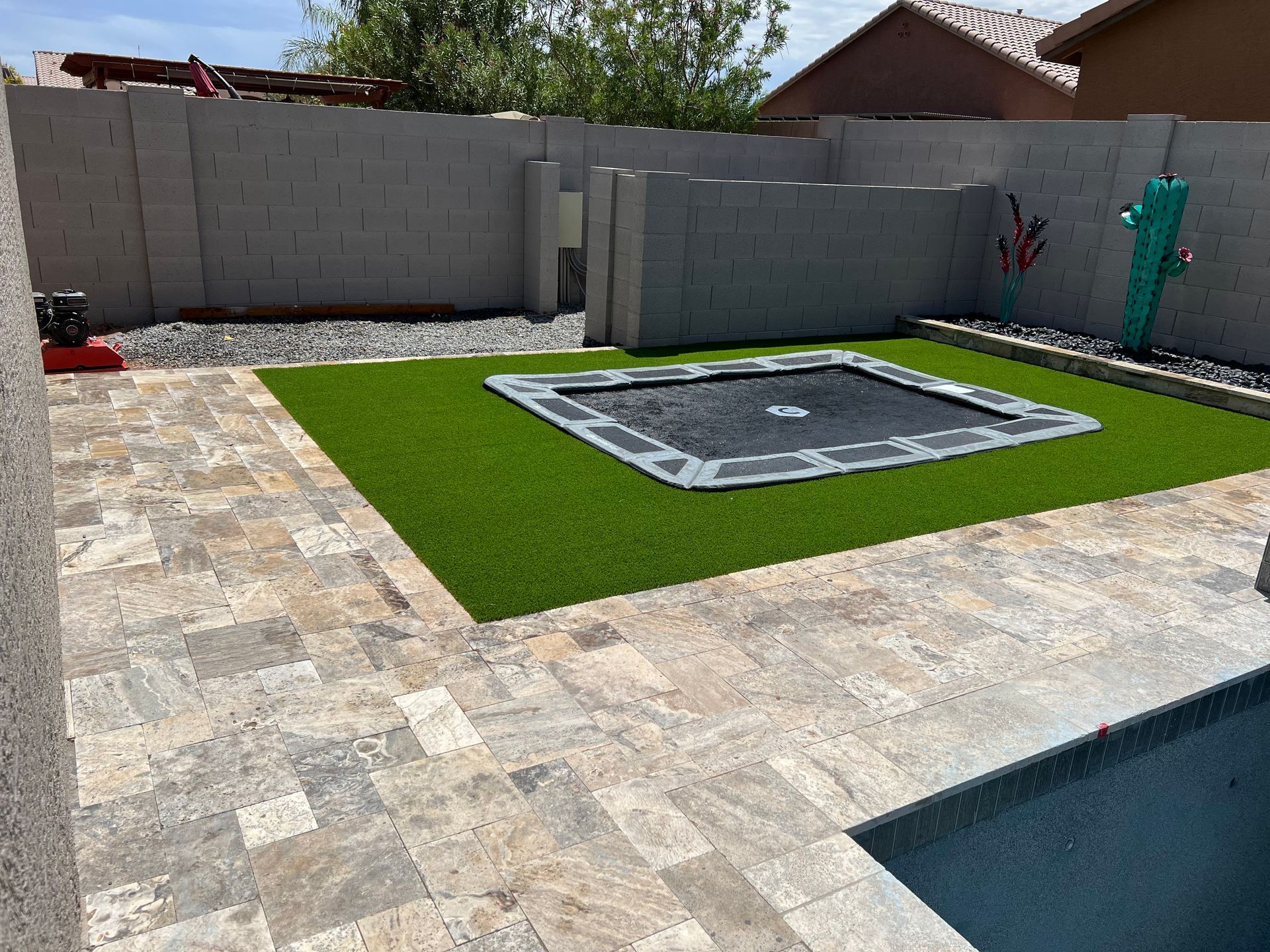 A backyard with a rectangular in-ground trampoline on artificial grass, surrounded by stone pavers and a pool.