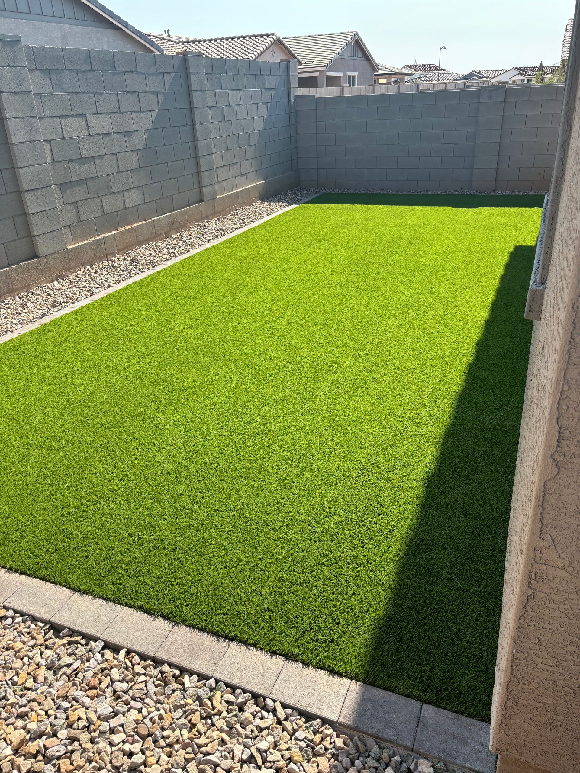 A backyard with vibrant green artificial turf, bordered by gravel and gray block walls.