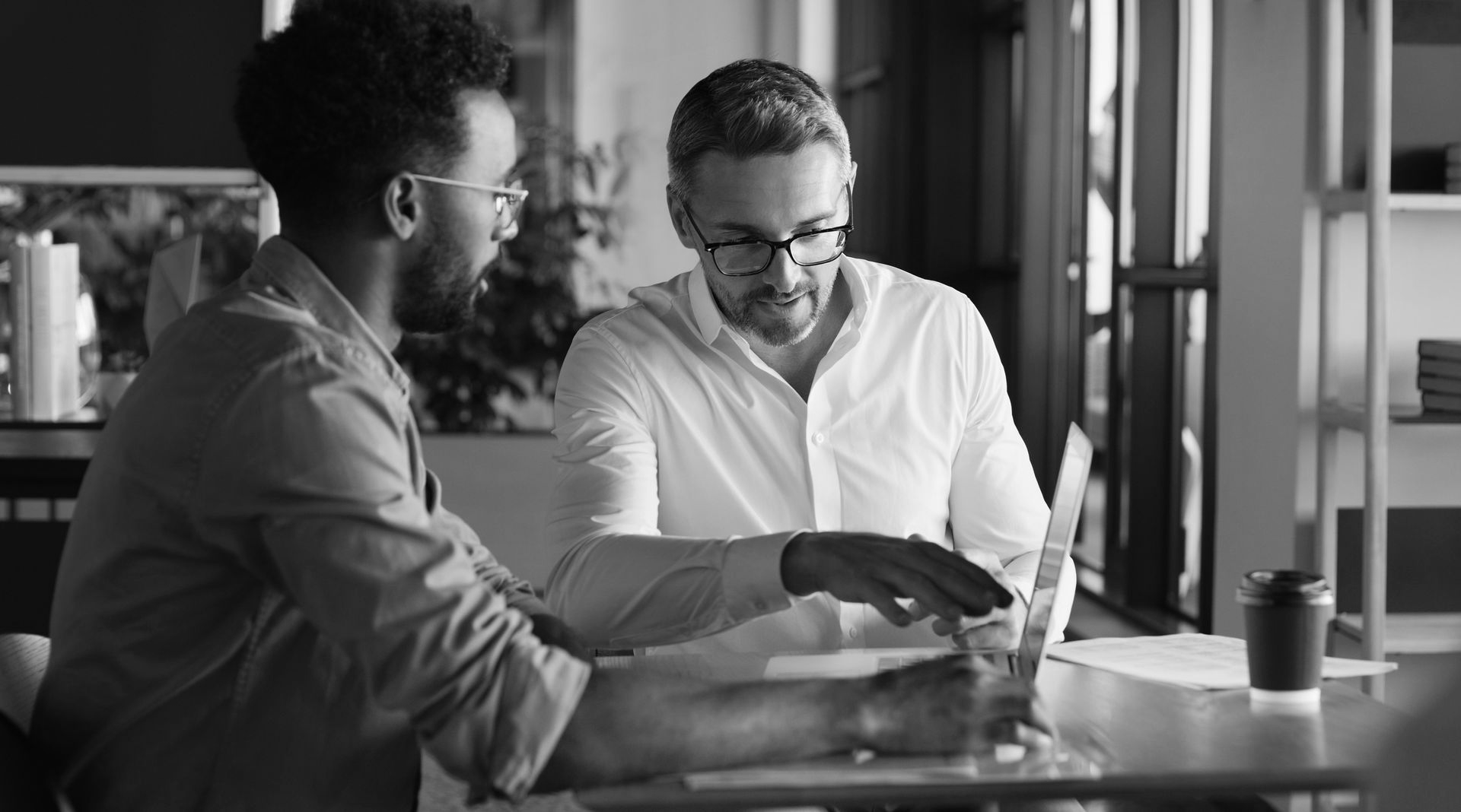 Two people working together at a table, one pointing at a laptop screen while the other looks on.