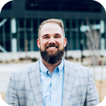 A smiling man with a beard wearing a light blue button-down shirt and a grey plaid blazer outdoors.