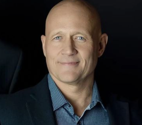 A person with a shaved head wearing a blue patterned button-down shirt and a dark blazer, smiling against a black background.