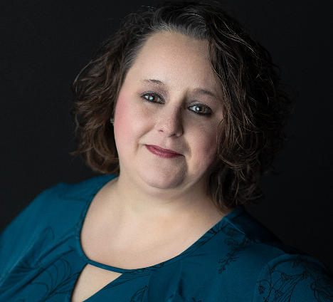 A person with shoulder-length curly brown hair wearing a teal top against a solid black background.