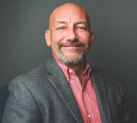 A man with a goatee and short hair wearing a grey blazer and red patterned shirt, smiling against a dark grey background.