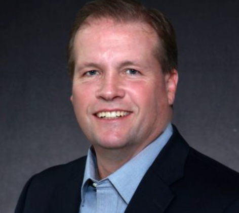 A smiling person in a blue button-down shirt and dark blazer against a dark gray background.