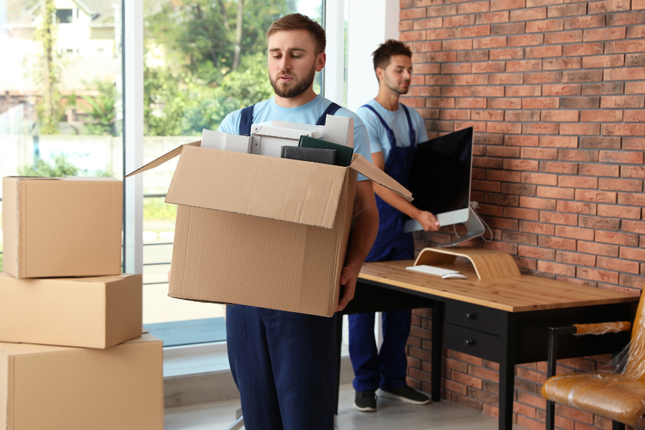 Two men are carrying boxes and a computer in a room.