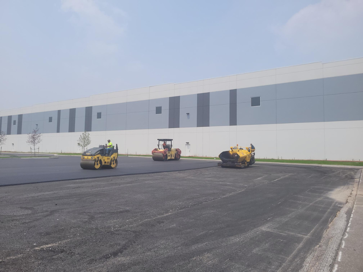 Three asphalt rollers are working on a parking lot in front of a large building.