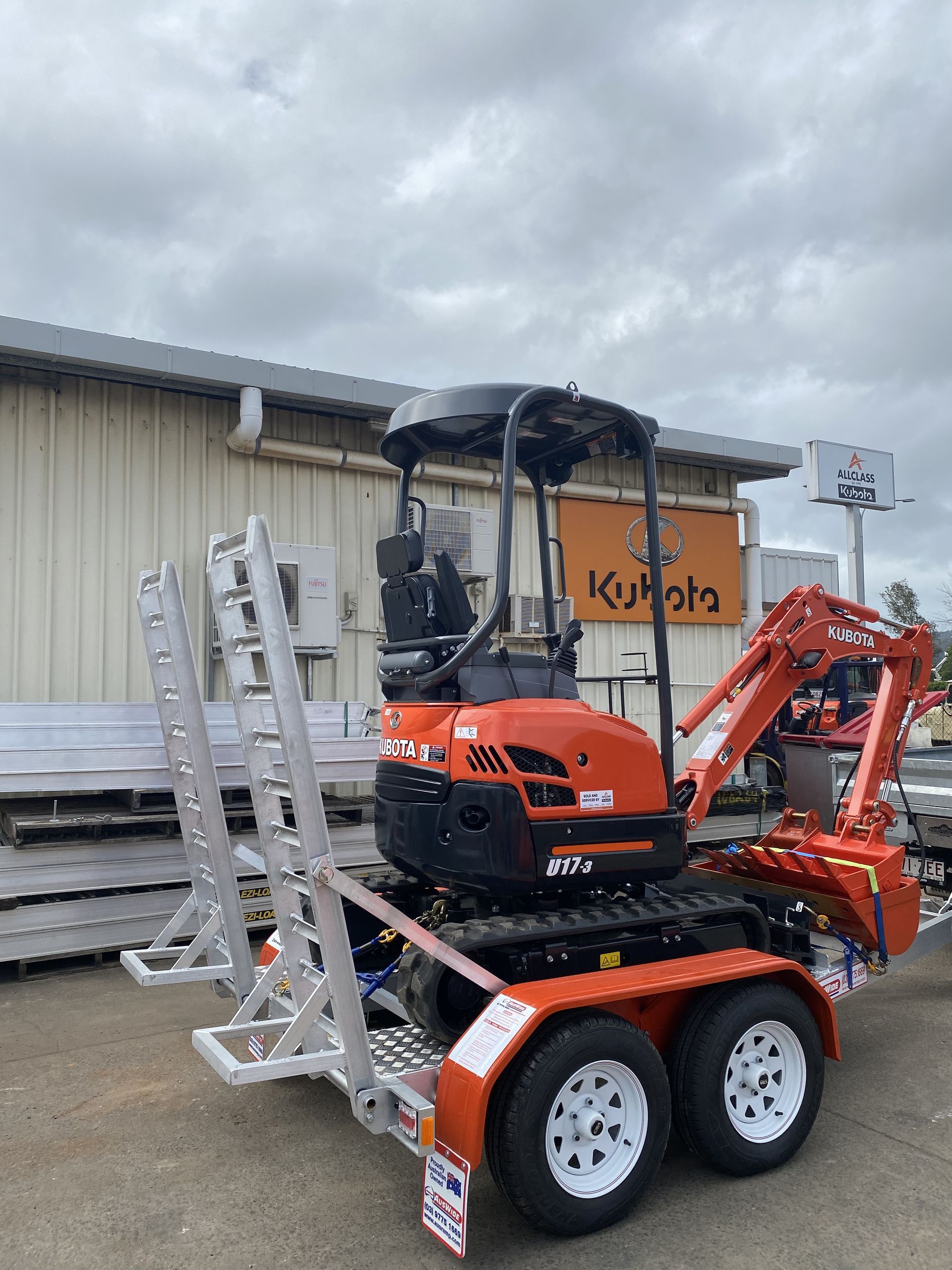 Orange Kubota excavator on a trailer with white wheels, parked in front of a building with a Kubota sign.