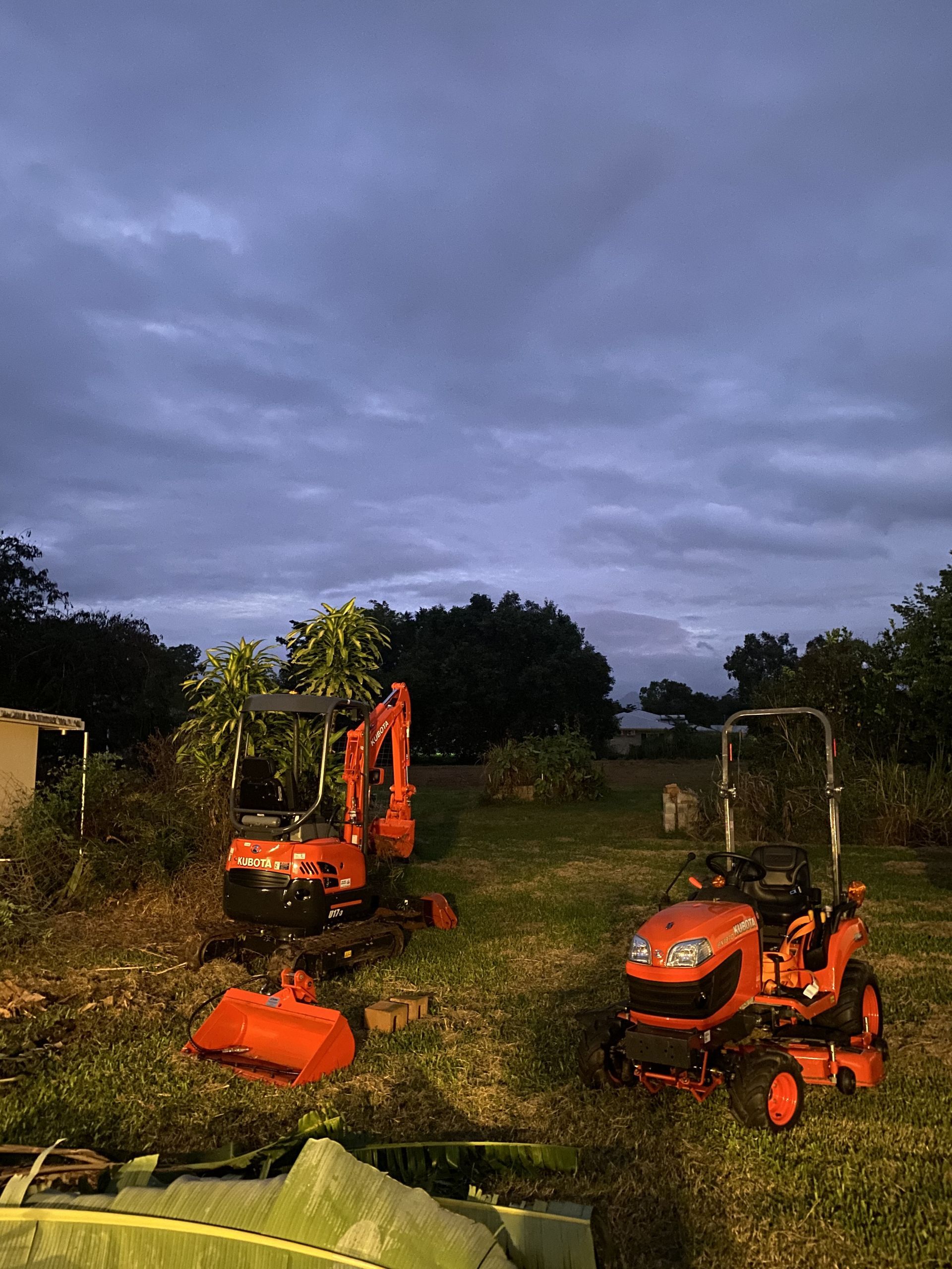 Orange excavator and riding mower on grassy field under overcast sky.