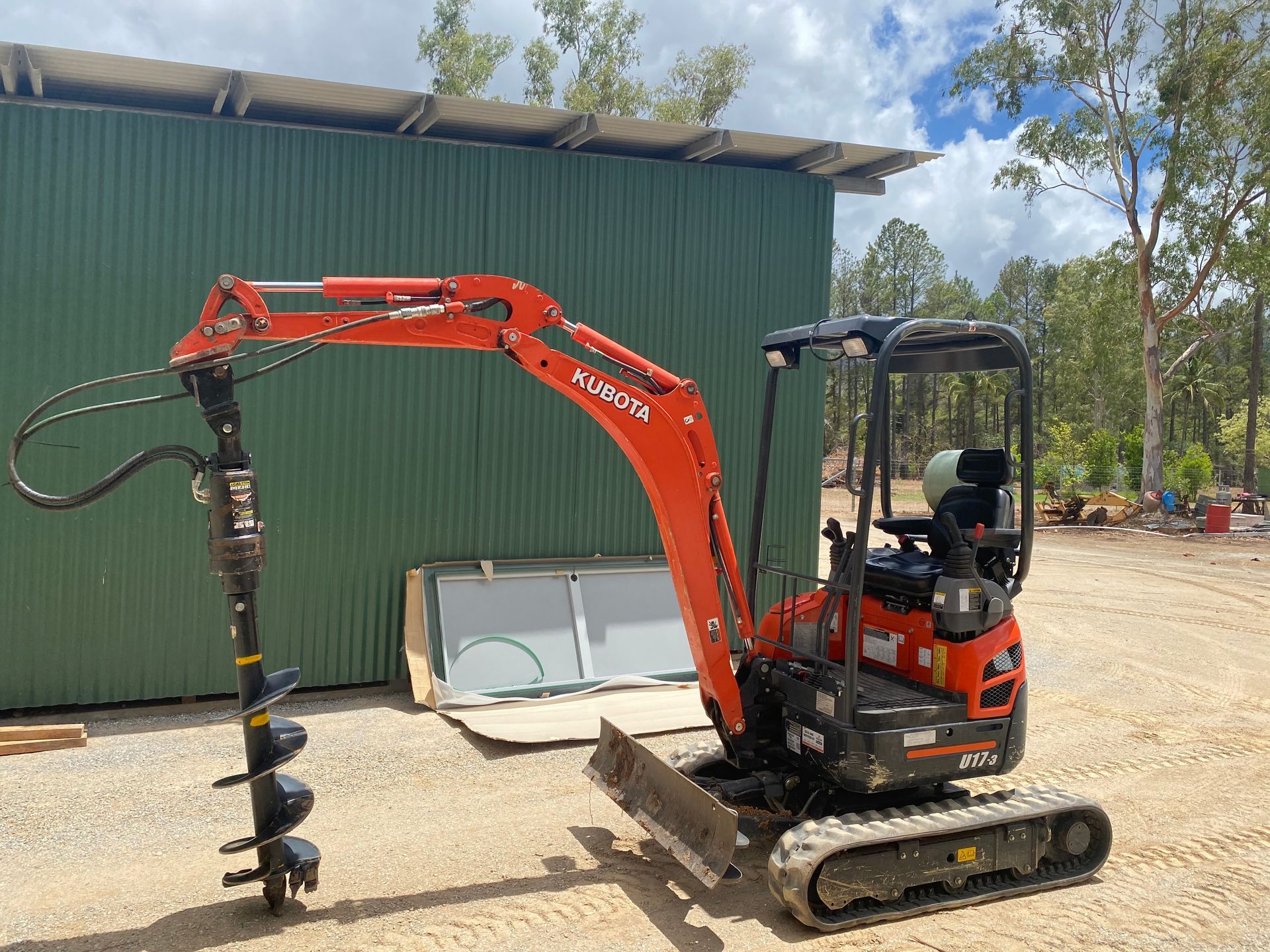 Orange Kubota mini excavator with auger attachment, parked in front of a green shed.— Mini Excavator Hire in Cairns, QLD