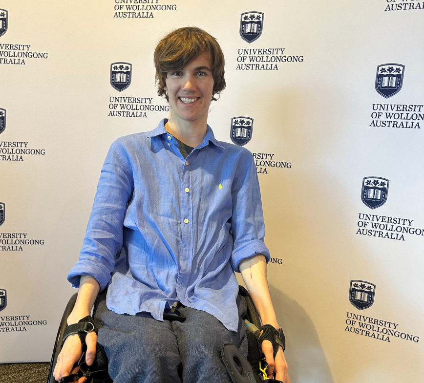 Hayden is a wheelchair user who smiles to the camera in front of a Wollongong University media wall. The wall shows the universities logo and blue text that reads University of Wollongong Australia.