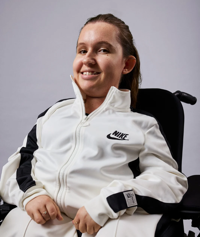 Close up studio portrait of Jamieson  in her electric wheelchair, wearing a white Nike matching tracksuit.