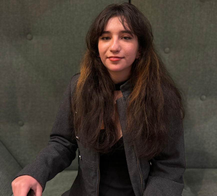 Close up photo of Jamie sitting on a green couch. She smiles to the camera with her long brown hair out, she holds a walking in one hand in front of her.