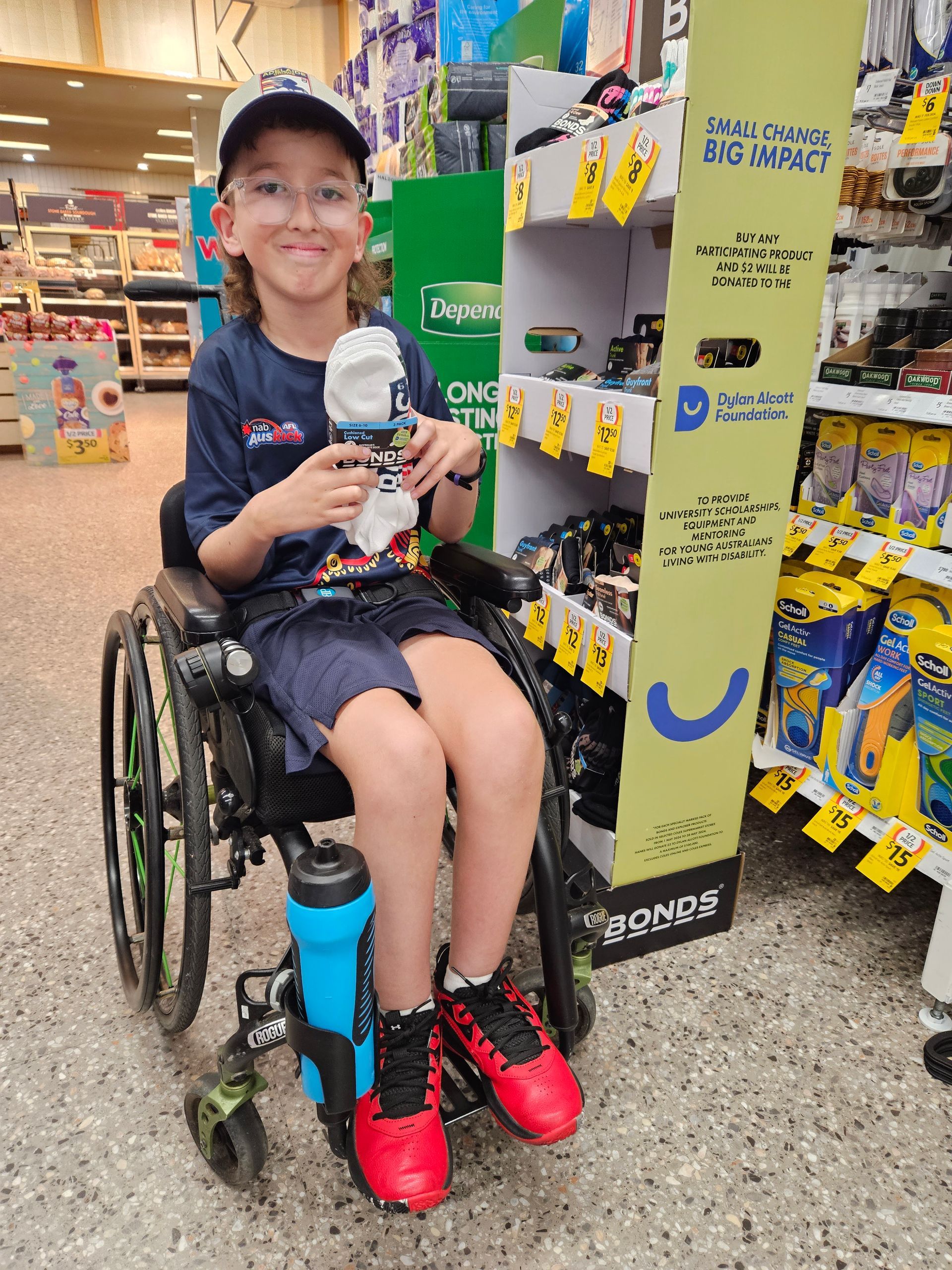 Recipient Alexander is a wheelchair user. He smiles to camera in aisle at a Coles store holding Bonds socks. The Bonds and Dylan Alcott Foundation branded product stand is to his right.
