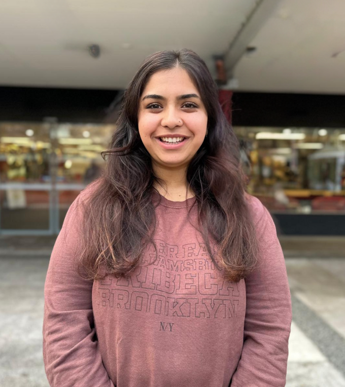Samiksha smiles to camera out the front of the library university which appears blurred. She wears a burgundy jumper.
