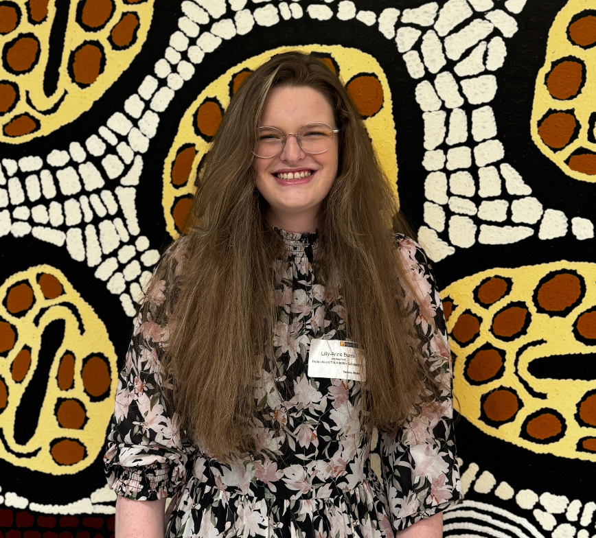 Lily-Anne wears glasses and her hair down. She smiles to the camera standing in front of a neutral coloured Indigenous piece of art work. She wears a name badge.