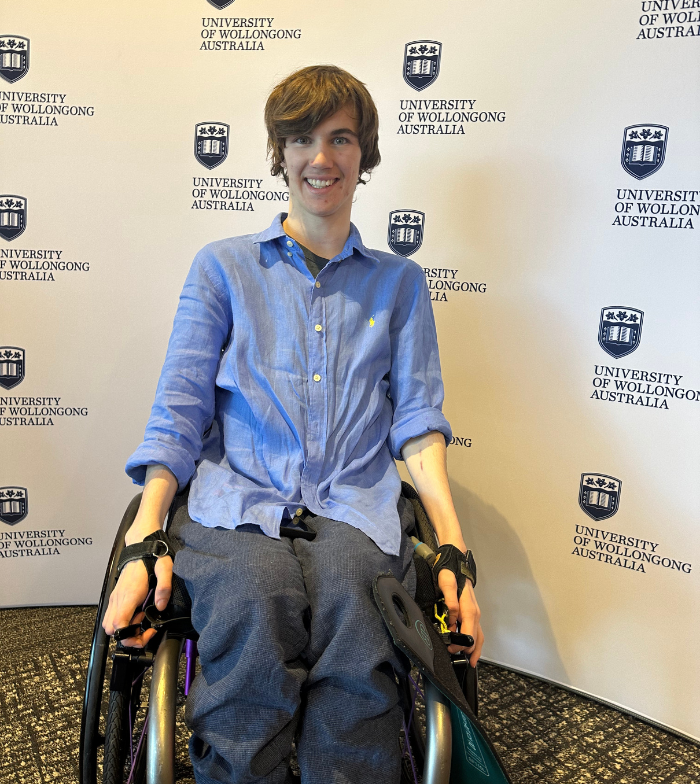 Hayden is a wheelchair user who smiles to the camera in front of a Wollongong University media wall. The wall shows the universities logo and blue text that reads University of Wollongong Australia.