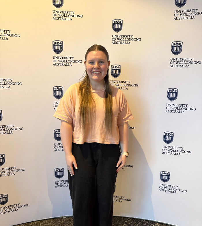Aleisha smiles to the camera with her hands by her sides. She stands in front of a Wollongong University media wall. The wall shows the universities logo and blue text that reads University of Wollongong Australia.