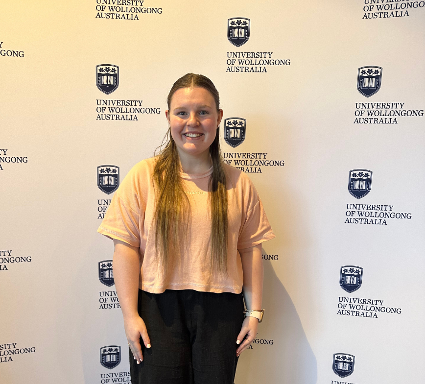 Aleisha smiles to the camera with her hands by her sides. She stands in front of a Wollongong University media wall. The wall shows the universities logo and blue text that reads University of Wollongong Australia.