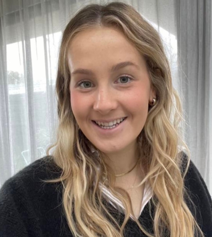 Close up photo of Mia smiling to camera. She has blonde hair and stands in front of a window with sheer curtains.