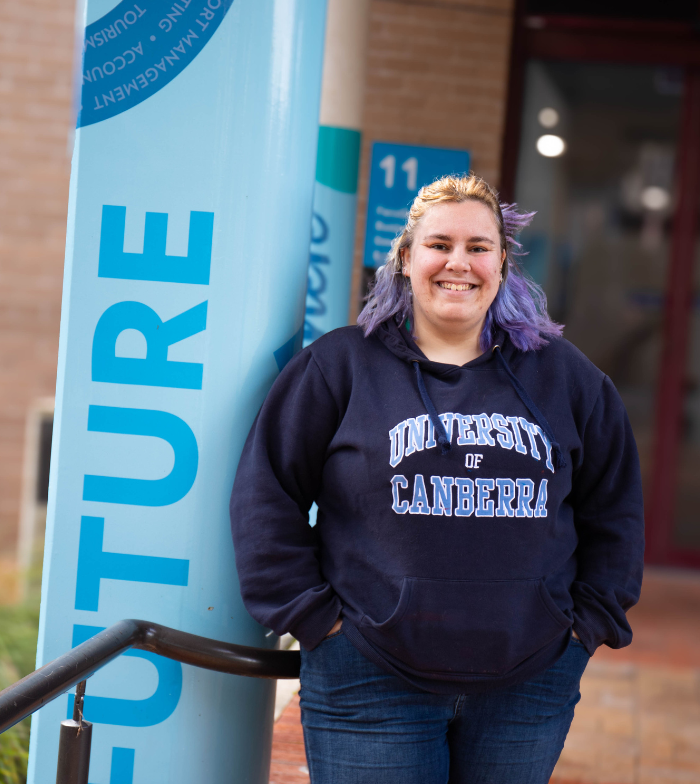 Jess leans against a blue pole on uni campus, hands in the pockets of a hoodie and smiling to camera.