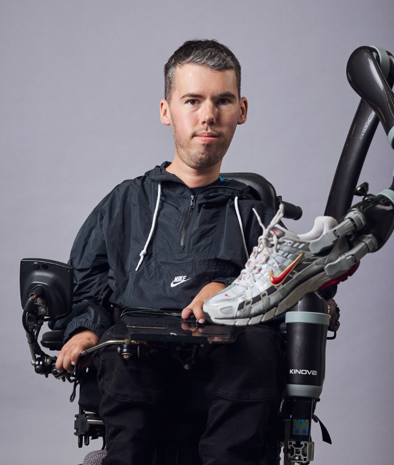 Studio portrait of Daniel in his electric wheelchair, wearing a black Nike spray. The mechanical arm of the chair is shown holding a Nike runner in front.
