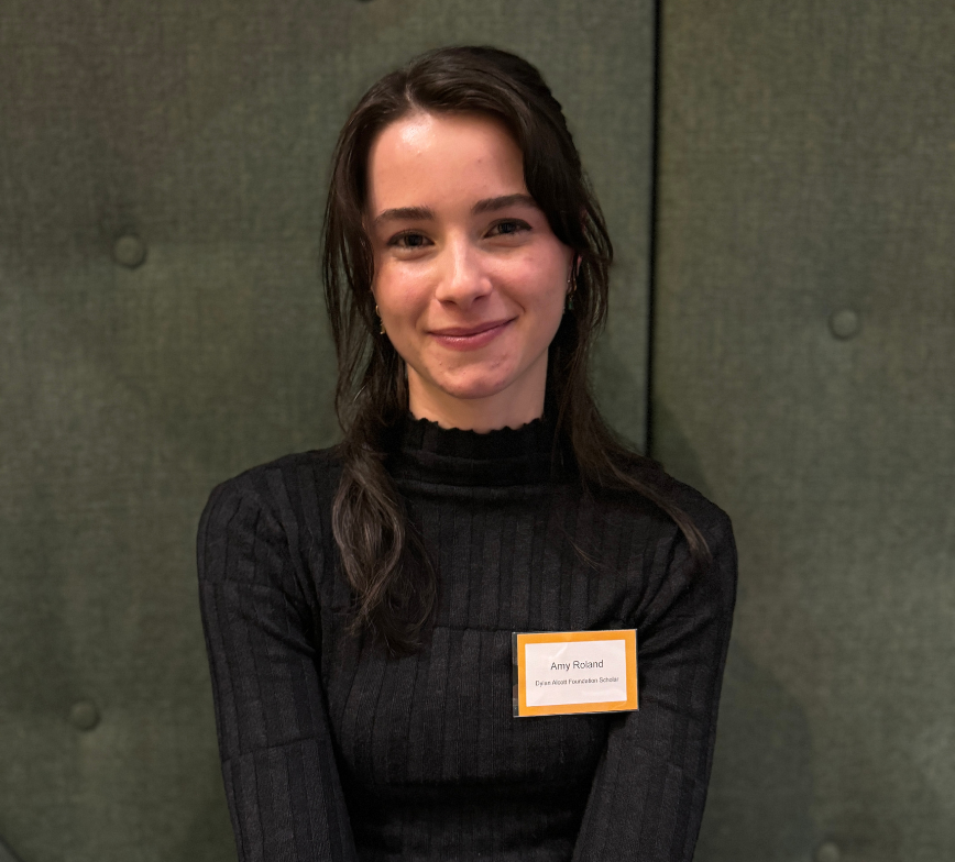 Close up shot of Amy sitting on a dark green couch smiling to camera. She has long black hair and wears a black long sleeve. A white and yellow name badge is pinned to her shirt. 