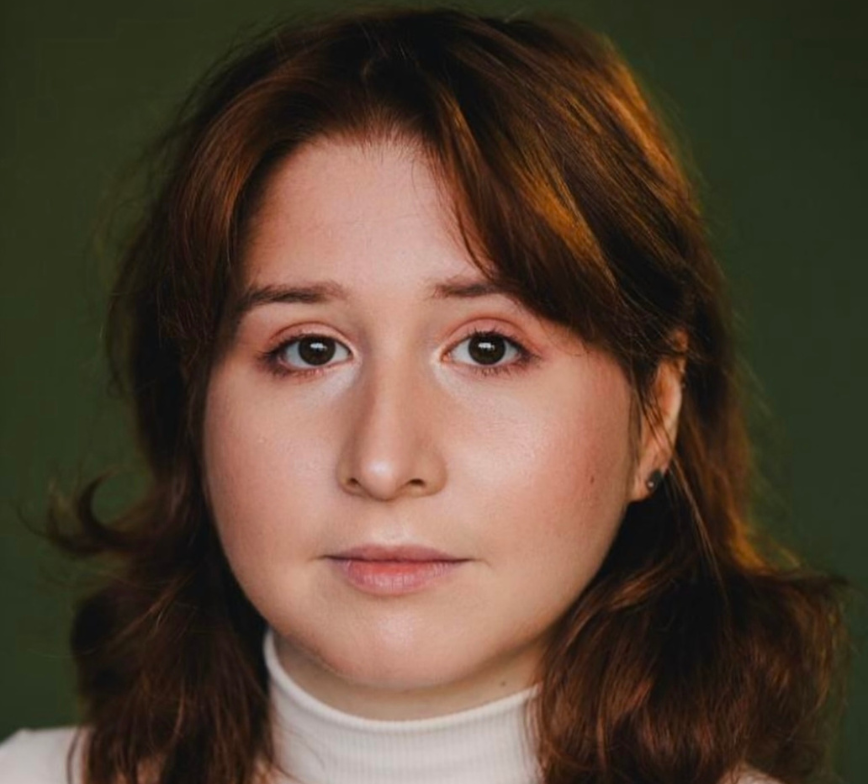 Close up photo of Rachel, she wears a white turtle neck and has browny red hair. The background is dark green. 