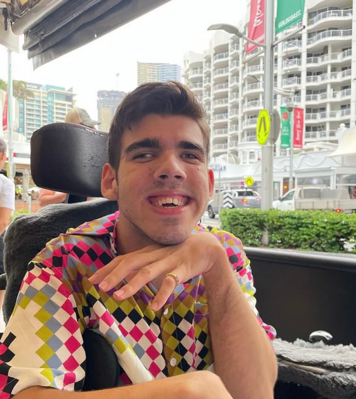 Max uses an electric wheelchair for mobility. He wears a vibrant checkered shirt and smiles to camera with one hand propped under his chin. The background shows white high rise buildings.