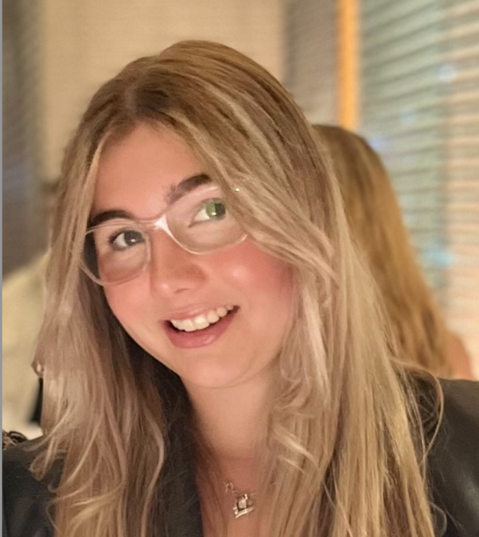 Close up photo of Sienna, she tilts her head to she side and smiles at the camera. She wears clear glasses and the background is blurred.