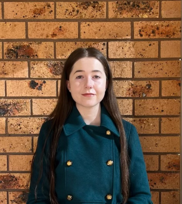 Charlotte stands facing the camera in a green double breasted jacket against a brown brick wall.
