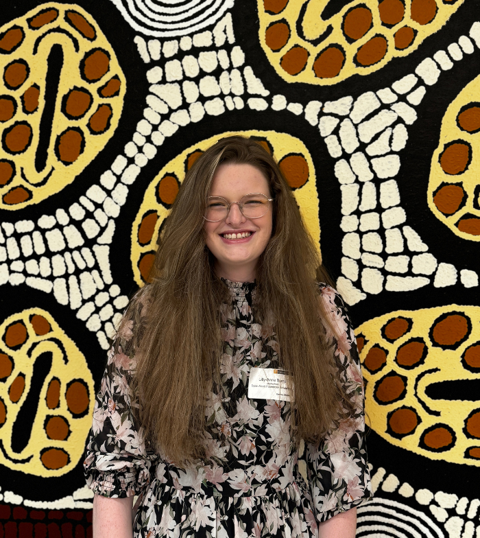 Lily-Anne wears glasses and her hair down. She smiles to the camera standing in front of a neutral coloured Indigenous piece of art work. She wears a name badge.