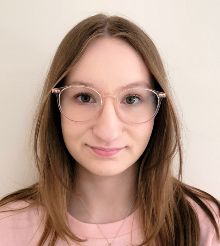 Close up shot of Michala front on smiling to camera. She wears light pink glasses and has light brown hair. The background is a white wall.