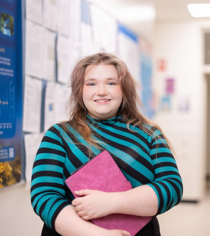 Daphne pictured holding books to chest as she smiles to camera in a university hallway that's blurred in the background.