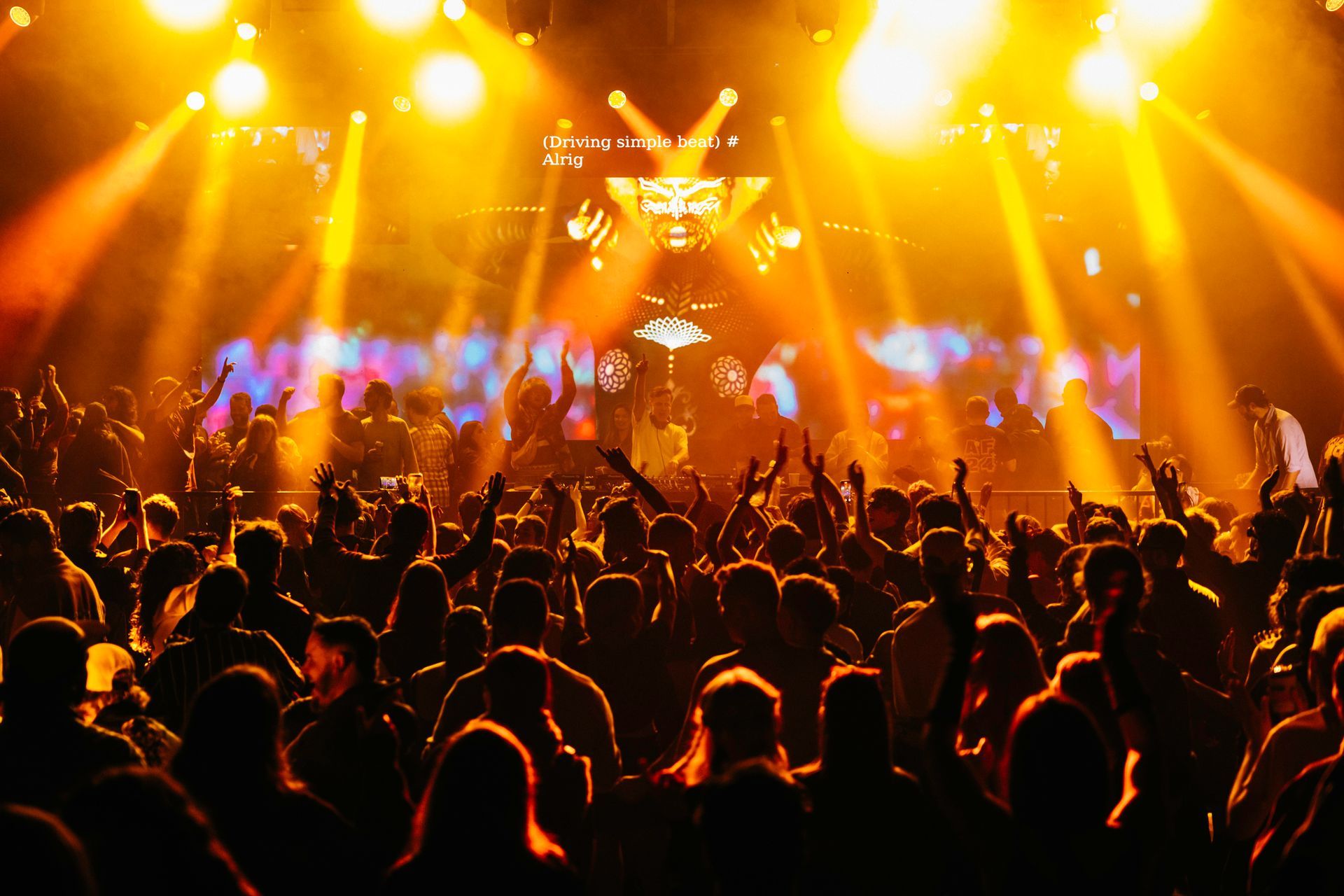 Inside a warehouse, a large crowd dances and raised their hands toward the stage where a DJ, PNAU is playing and dancing. Orange spotlights shine from the roof with orange visuals on the main screen behind the DJ and crowd on platform.
