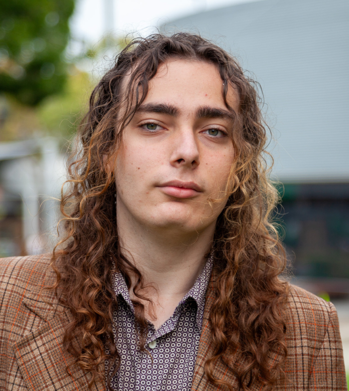 Pictured outdoors with a blurred background, Saul has long brown wavy hair and wears a brown tweed check jacket.