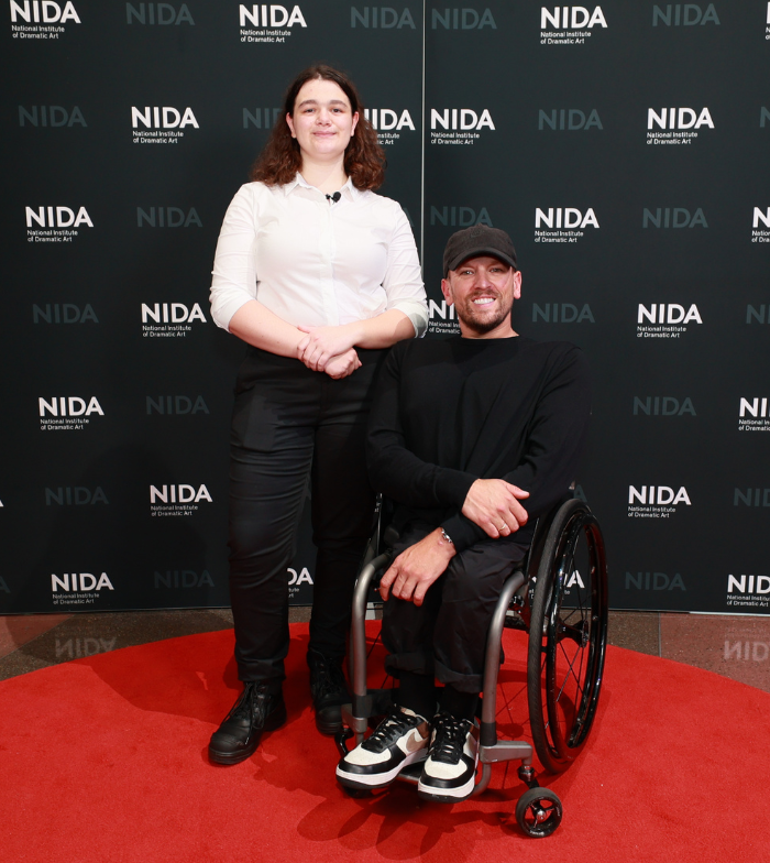 Kaia (left) stands next to Dylan (right) who is a wheelchair user. They smile to camera on red carpet with a black and white NIDA media wall behind them.