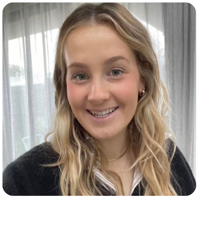 Close up photo of Mia smiling to camera. She has blonde hair and stands in front of a window with sheer curtains.