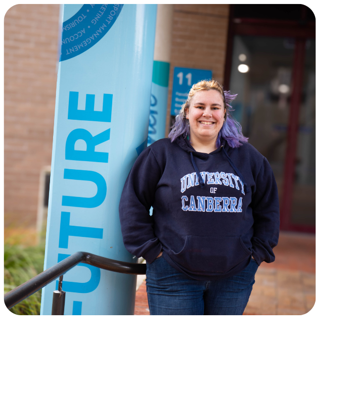 Jess leans against a blue pole on uni campus, hands in the pockets of a hoodie and smiling to camera.